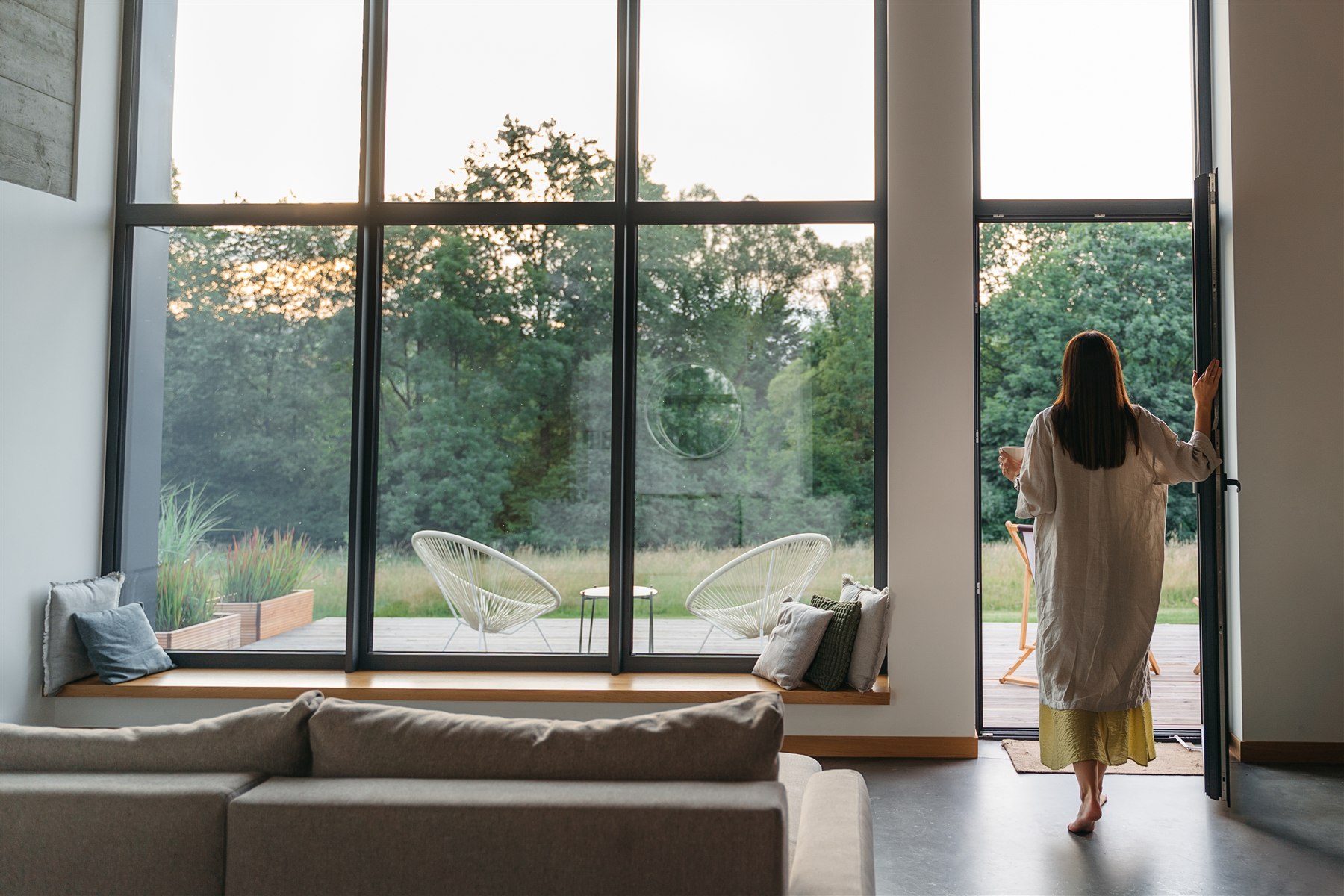 A woman standing quietly beside a large window at home.