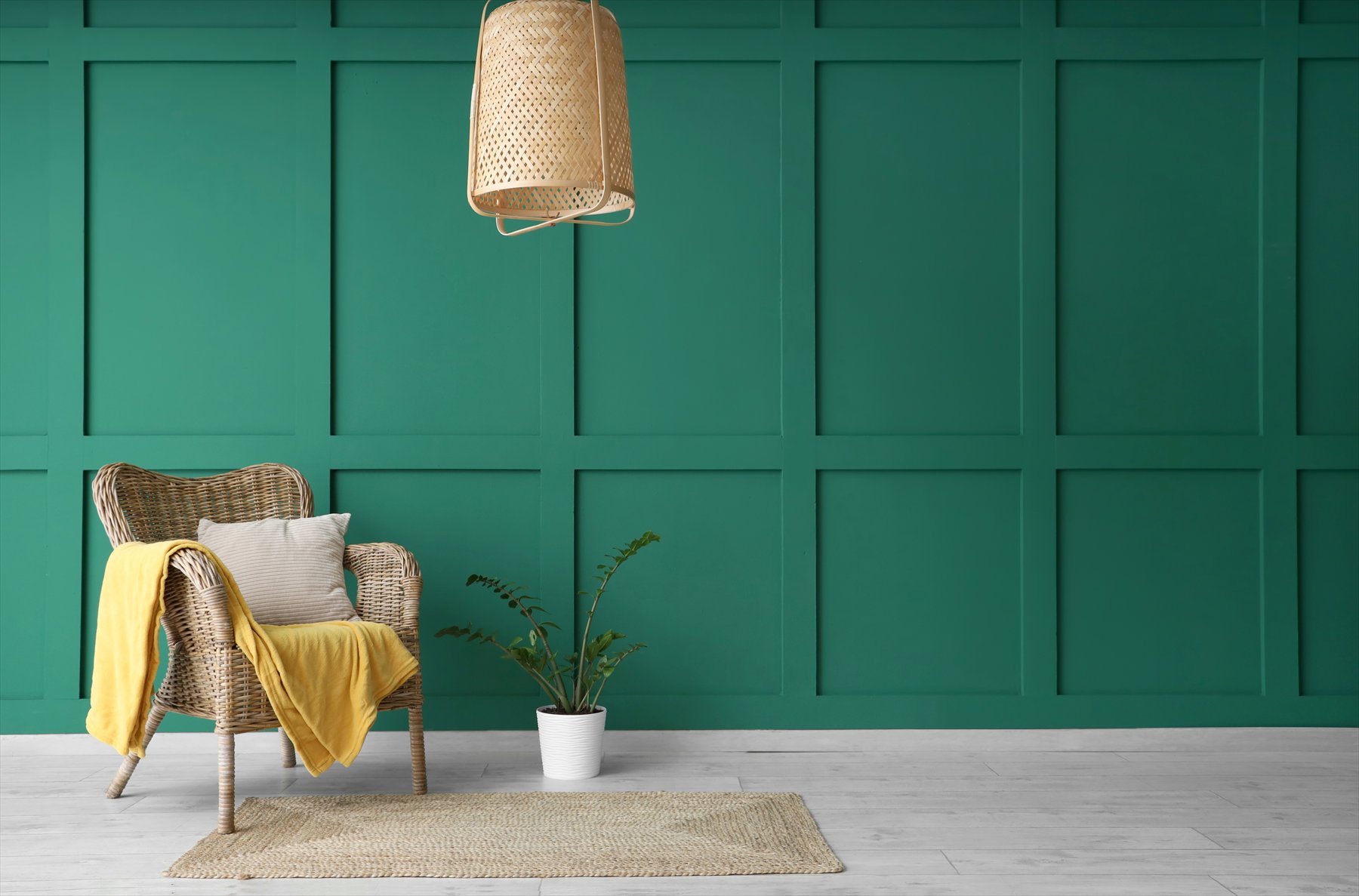 A wicker chair with a yellow throw beside a plant against a teal wall.