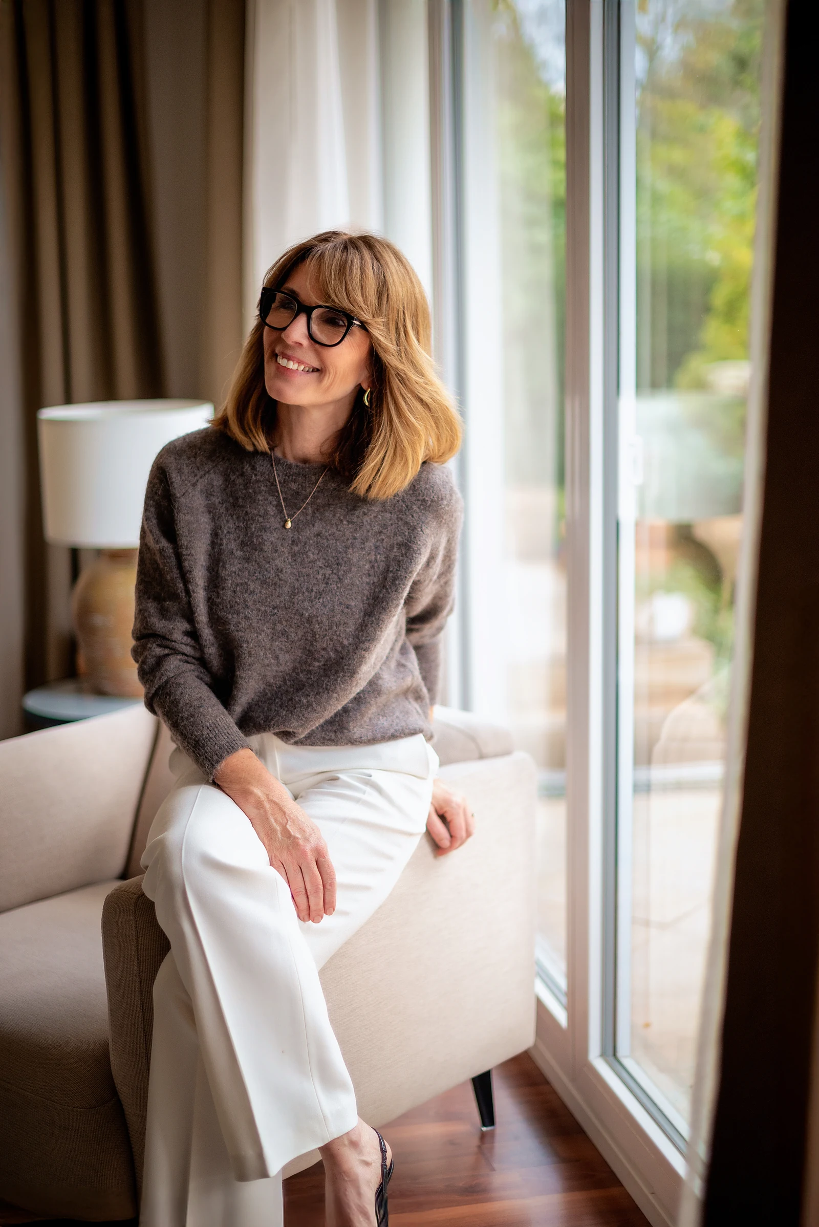 A woman in her fifties sits on the arm of a chair by a tall window at home, smiling.