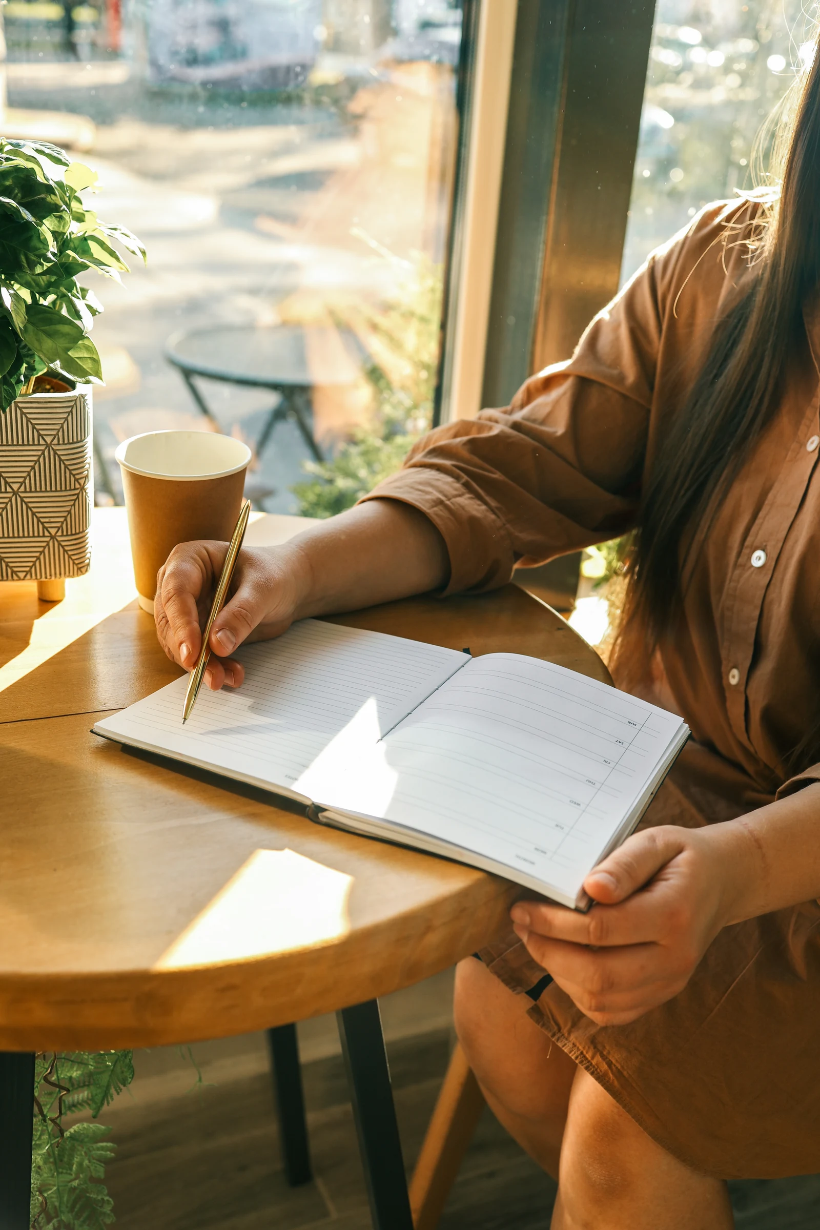 A woman at a sunlit café table writes in a dated planner with a gold pen.